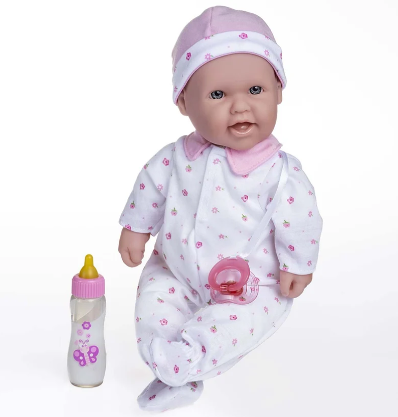 A baby doll wearing a white footed onesie and matching hat with pink floral prints, posed next to a small pink-and-yellow baby bottle on a white background.
